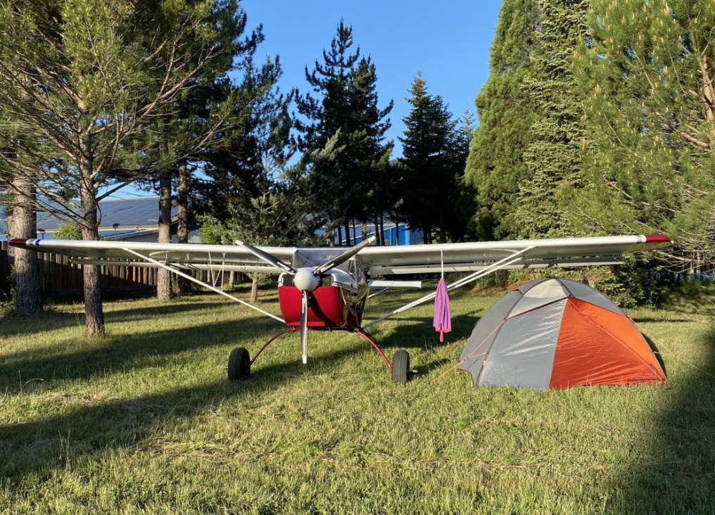 Photo d'une tente dans un jardin, sous l'aile d'un avion au sol. 